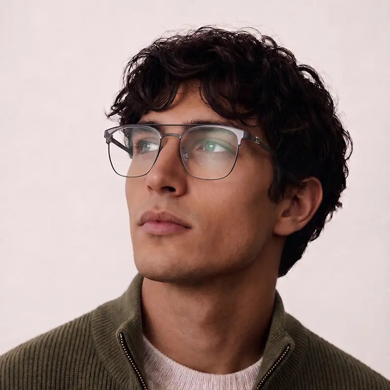 A man with curly dark hair wearing browline pure titanium designer glasses frame with clear lenses, shown in a close-up portrait against a plain light background.