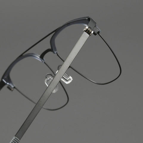 Close-up of a pair of browline designer glasses frames with a slim titanium gunmetal frame and thin black pure titanium temples, shown against a neutral gray background.