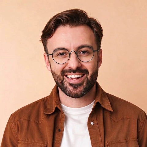 A smiling man with a beard and Round Black Full Rim Titanium Eyeglasses, wearing a brown overshirt over a white t-shirt, set against a solid beige background.