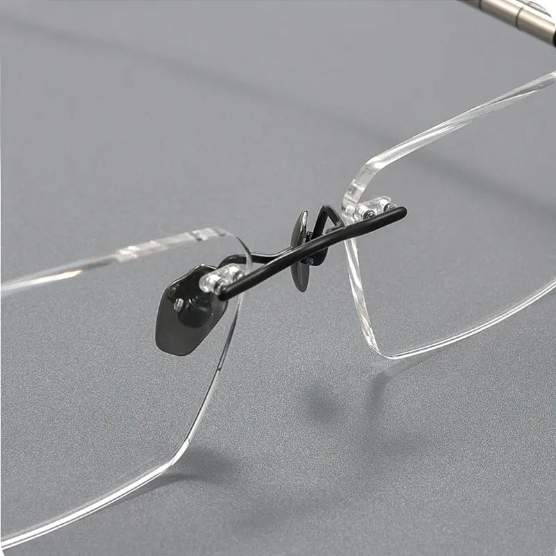Close-up of a pair of rimless eyeglasses with clear transparent frames and black titanium nose pads on a gray background.
