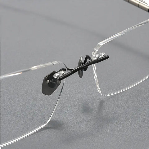 Close-up of a pair of rimless eyeglasses with clear transparent frames and black titanium nose pads on a gray background.