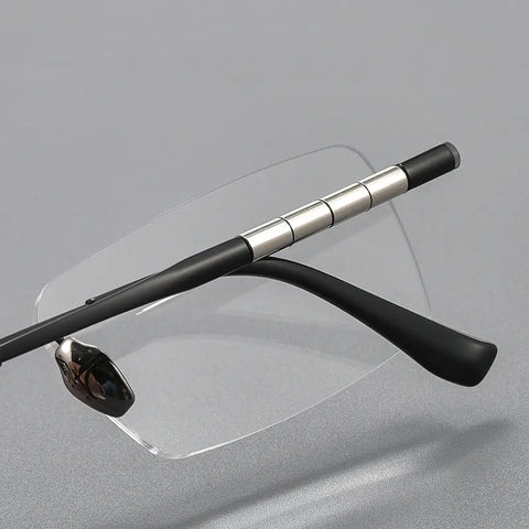 A pair of designer rimless eyeglasses with a clear lens, black temples, and silver metal hinge accents, displayed on a neutral gray background.