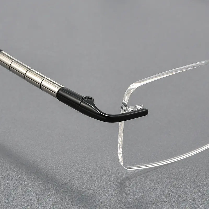Close-up of a clear rimless eyeglass lens meeting a silver metal temple with a black nose bridge on a gray background.