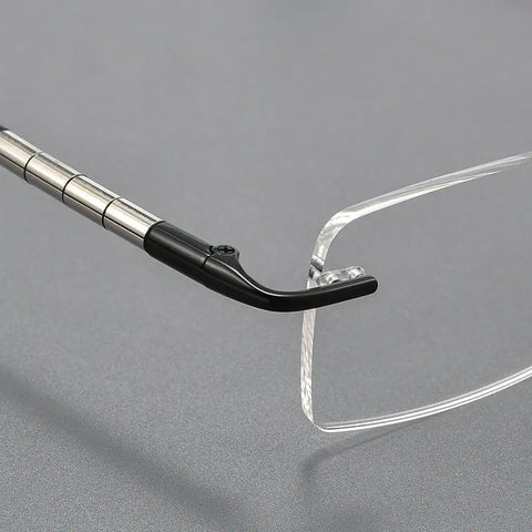 Close-up of a clear rimless eyeglass lens meeting a silver metal temple with a black nose bridge on a gray background.