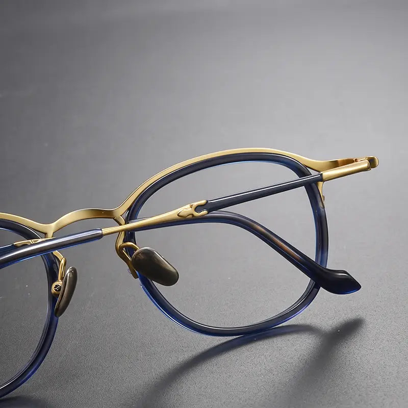 Close-up of a pair of pure titanium vintage designer eyelasses from Verhalo Vision with a gold titanium brow bar and bridge, paired with translucent dark blue acetate rims and matching blue temple tips, resting on a dark gray surface.