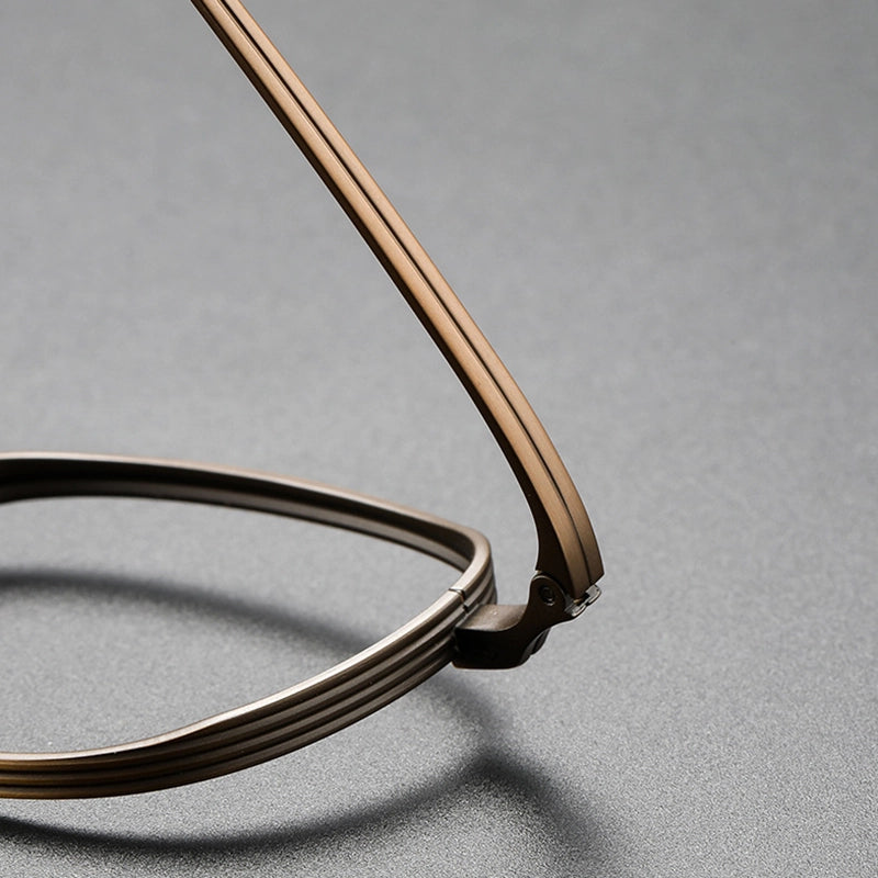 Close-up of a pair of round pure titanium eyeglasses with a thin, multi-strand metal frame in a warm bronze finish, resting on a textured gray surface.