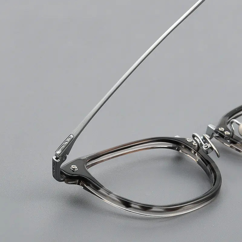Close-up of a pair of thin titanium eyeglasses with a hinge design, showing the temple arm and part of the frame on a smooth gray surface.