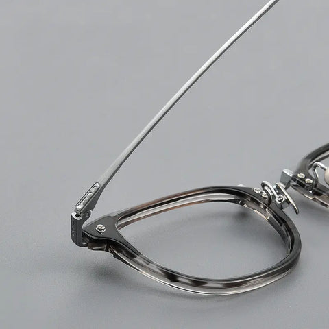 Close-up of a pair of thin titanium eyeglasses with a hinge design, showing the temple arm and part of the frame on a smooth gray surface.