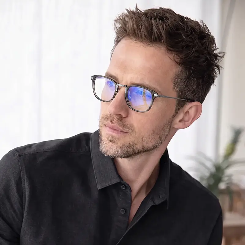 A man wearing round designer titanium eyeglasses with blue light lenses, dressed in a black button-up shirt against a bright, softly lit indoor background.
