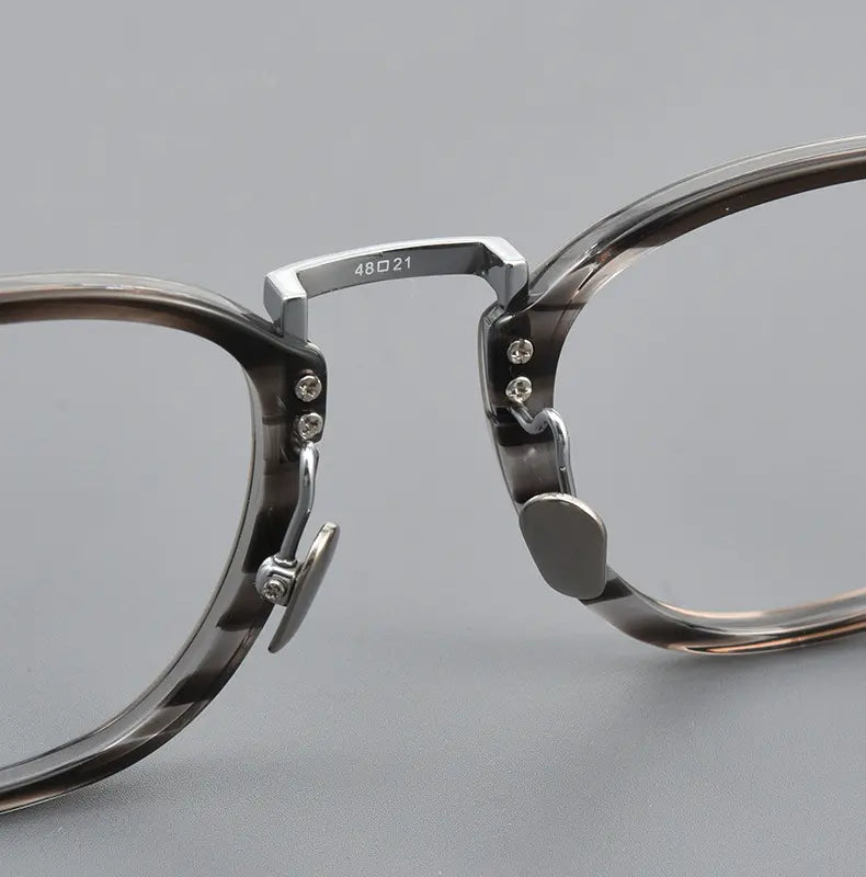 Close-up of a pair of translucent smoky gray acetate and titanium eyeglass frames with silver titanium hinge hardware, shown on a neutral gray background.