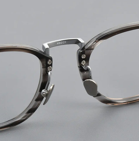 Close-up of a pair of translucent smoky gray acetate and titanium eyeglass frames with silver titanium hinge hardware, shown on a neutral gray background.