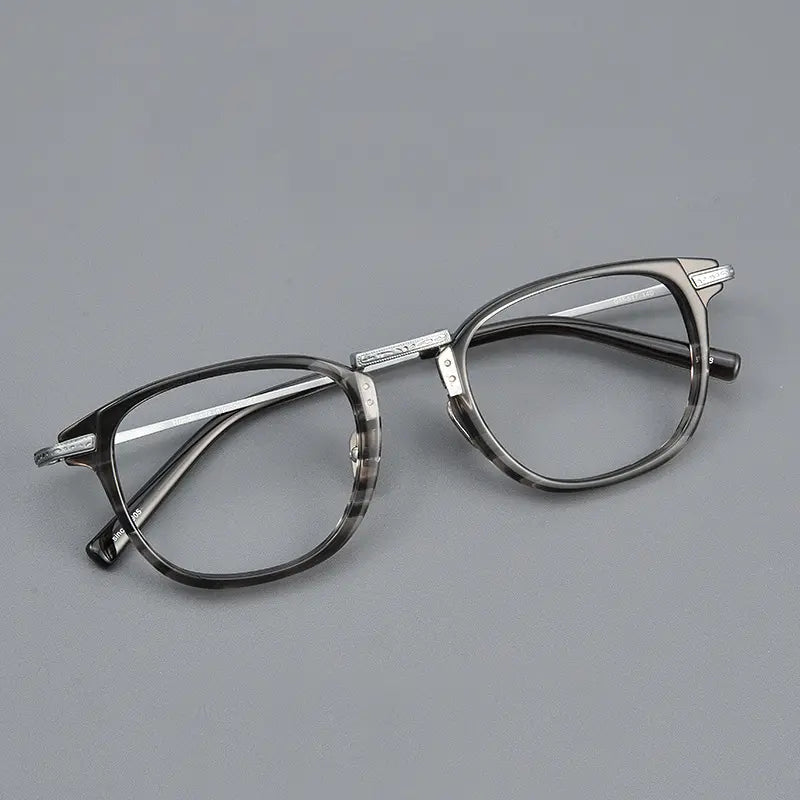 A pair of designer titanium eyeglasses with a dark gray and black marbled acetate front frame, silver titanium bridge and temples, and clear lenses, shown on a plain gay background.
