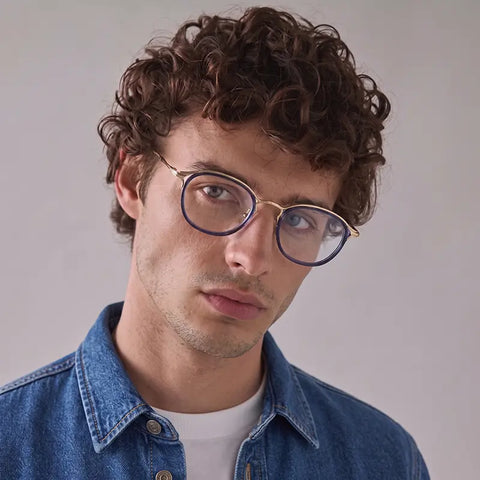 A close-up portrait of a man with curly brown hair wearing round Vintage Designer Eyelasses from Verhalo Vision with a gold bridge and dark blue rims, paired with a blue denim shirt over a white undershirt, against a plain light gray background.