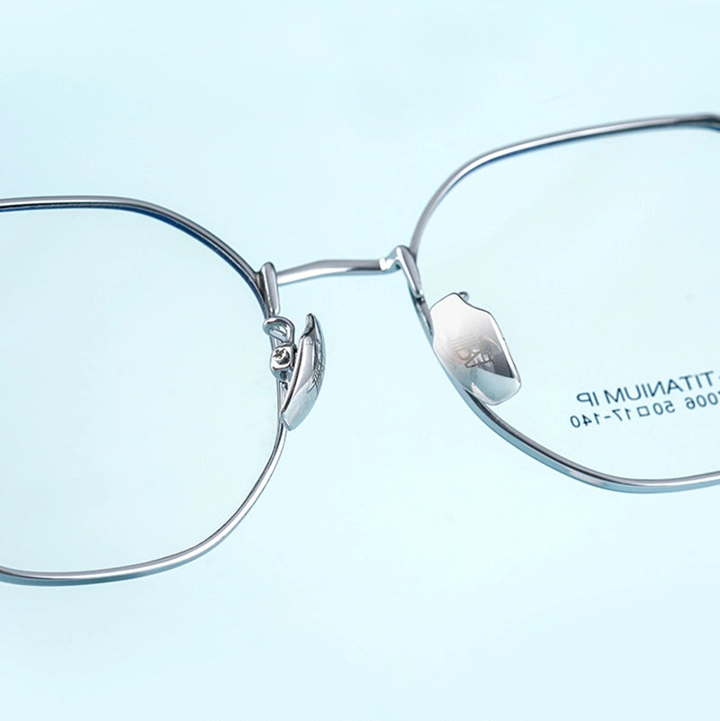 Close-up of silver titanium geometric glasses frame from Verhalo Vision with clear lenses on a pale blue background. The right lens shows printed text reading 'TITANIUM IP'.