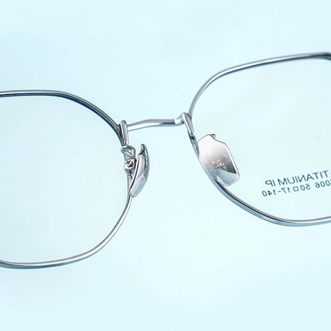 Close-up of silver titanium geometric glasses frame from Verhalo Vision with clear lenses on a pale blue background. The right lens shows printed text reading 'TITANIUM IP'.