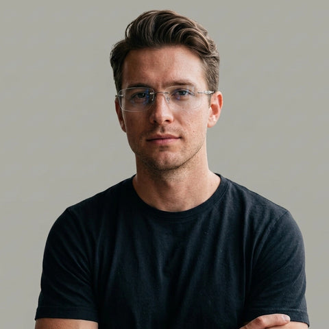 A man with styled brown hair and minimalist silver titanium rimless spectacles for Men, wearing a dark navy blue t-shirt, photographed against a solid light gray background.