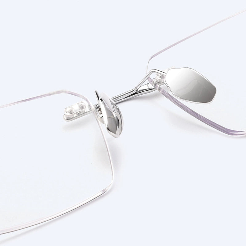 Close-up of a pair of minimalist silver titanium rimless spectacles for Men with a silver titanium hinge and clear lenses on a light gray background.
