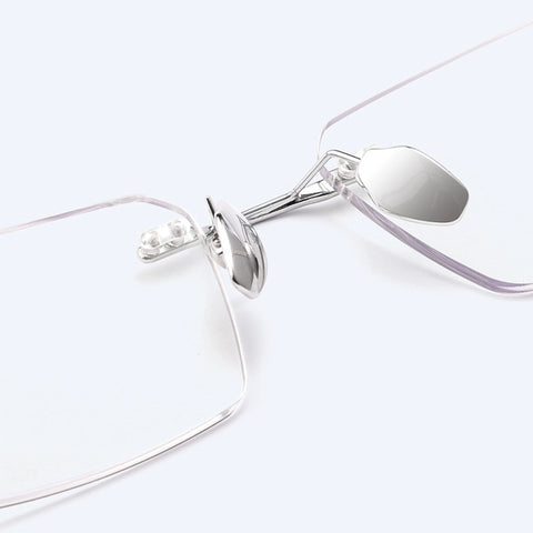 Close-up of a pair of minimalist silver titanium rimless spectacles for Men with a silver titanium hinge and clear lenses on a light gray background.
