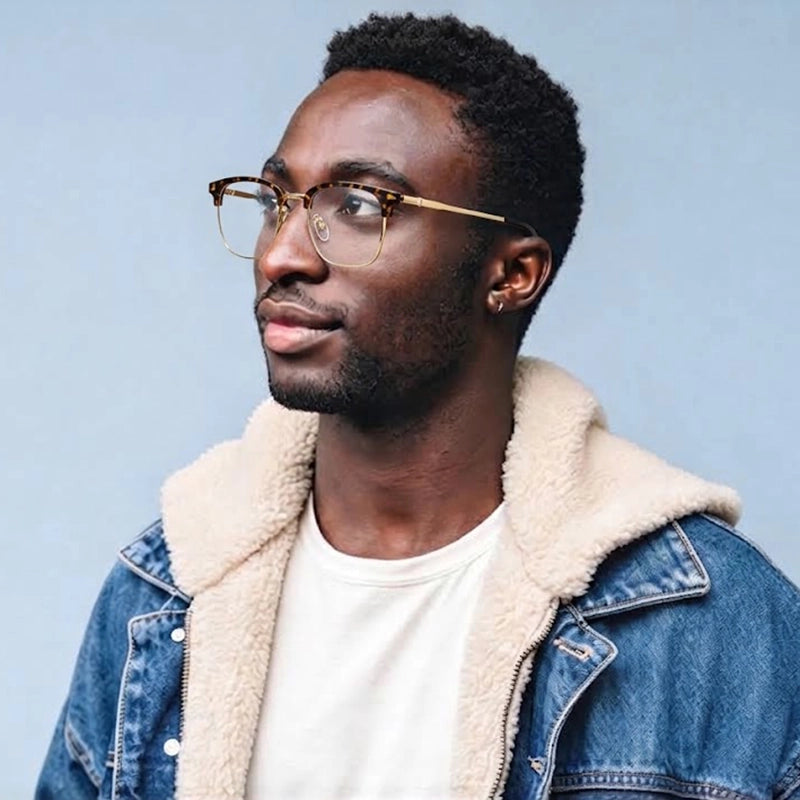 A man wearing tortoiseshell Browline Designer Pure Titanium Eyeglasses with a gold bridge, styled with a blue denim jacket featuring a beige sherpa lining over a white t-shirt, set against a solid light blue background.