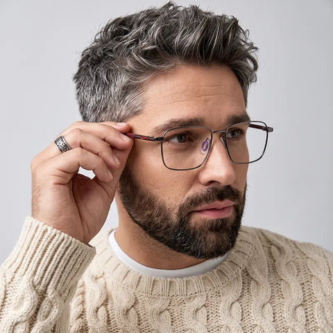 A close-up of a man with gray hair and a beard adjusting his pure titanium designer eyewear, wearing a cream cable-knit sweater against a light gray background.