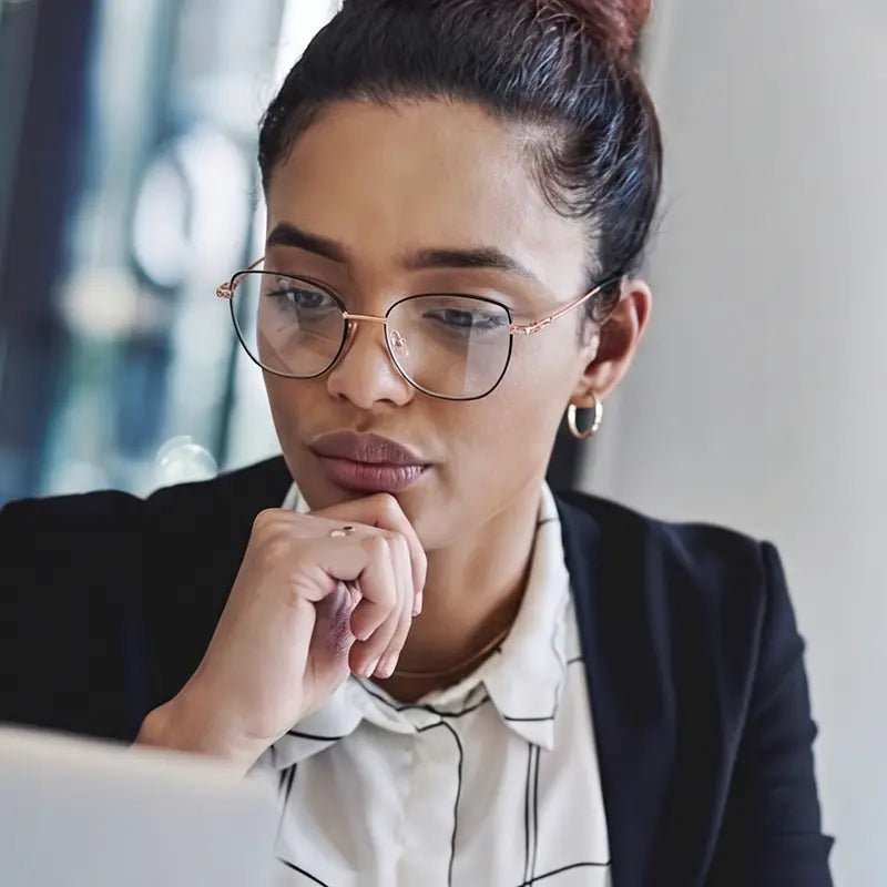 A woman with black and gold cat eye full-rim pure titanium eyeglasses from Verhalovision, a black blazer, and a white collared shirt looking down thoughtfully. She is wearing rose gold hoop earrings and a gold chain necklace.